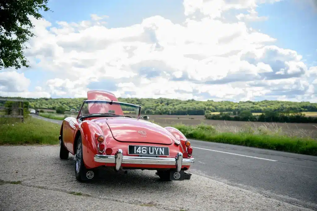 Un MG clásico, probablemente un MGB de los 60/70, rojo, descapotable, matrícula “146 UYN”, estacionado en una carretera con campos verdes y cielo parcialmente nublado de fondo. Se le han estropeado las vacaciones de verano.