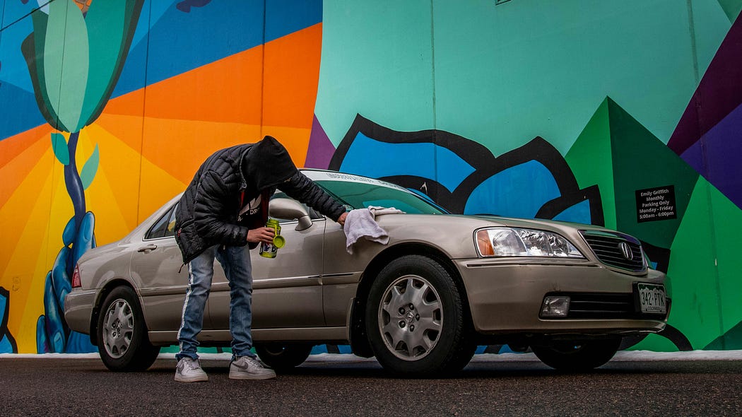 Comprar un coche de segunda mano: Un hombre limpia su coche con un trapo. El coche es de color marrón y es un modelo de en torno al año 2000. Hay un grafiti colorido tras el coche en el que destacan los colores naranja, azul, verde, y turquesa.