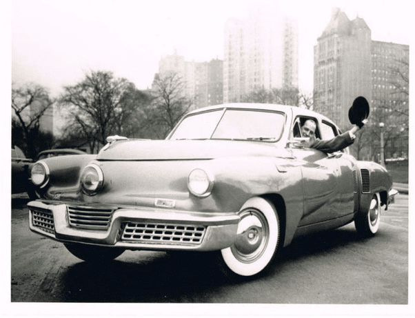 La imagen muestra una fotografía en blanco y negro de un coche antiguo con un diseño único, destacando parachoques delanteros prominentes y faros circulares. El coche está estacionado en lo que parece ser una carretera urbana con árboles y edificios altos en el fondo. Una persona está sentada en el asiento del conductor, extendiendo su brazo por la ventana, sosteniendo lo que parece ser un sombrero o un objeto redondo.