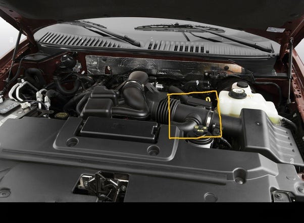 El vano motor de un Ford Expedition. Se ve el motor y los plásticos que lo protegen. Predomina el color negro de los plásticos, y hay un depósoto de anticongelante blanco translucido en el que se ve anticongelante amarillo. La imagen tiene marcado en amarillo el tubo de admisión del Ford Expedition.