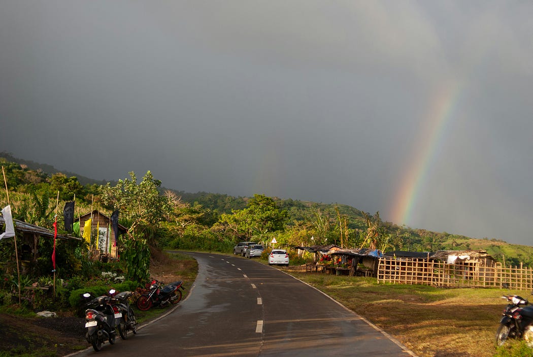 Una carretera rural despejada y húmeda. Hay nubes en el cielo y un arcoiris. También hay dos motos aparcadas en el lado izquierdo. Hay vegetación verde al fondo y parece que la foto está tomada en Asia.