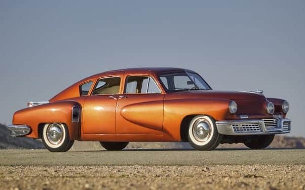 La imagen muestra un coche antiguo, de mediados del siglo XX, con un diseño elegante y una brillante pintura naranja. El vehículo está posicionado en lo que parece ser una carretera abierta o un área despejada. El diseño clásico del coche, como las formas redondeadas de la carrocería, los detalles cromados y los neumáticos con banda blanca, lo hacen interesante.