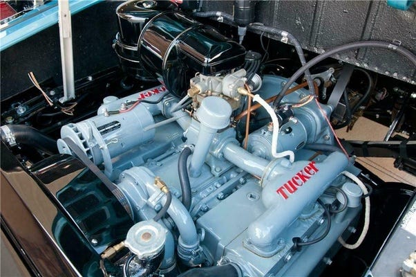 La imagen muestra una vista de cerca del motor de un coche antiguo, específicamente etiquetado como “TUCKER”. El motor parece estar bien mantenido y está instalado en el vehículo. Los componentes, incluyendo el filtro de aire, las tapas de las válvulas y varios tubos y conexiones, están pintados de un color azul claro uniforme con algunas partes en negro. La marca “TUCKER” se muestra de manera prominente en la tapa de las válvulas con letras rojas y un contorno blanco.