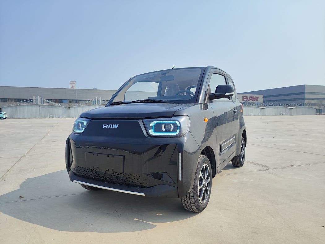 Un microcoche de color negro con luces LED. Está aparcado en una zona industrial con fábricas al fondo.