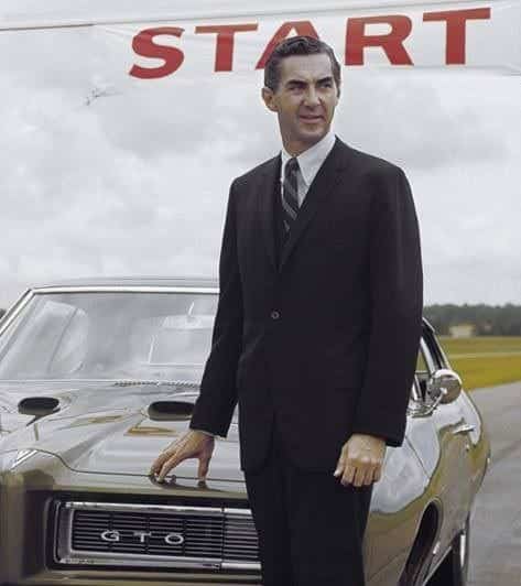 Entendido. Aquí tienes una descripción revisada de la imagen, sin mencionar la pixelación y resaltando la presencia de John DeLorean:
 
 La imagen muestra a un John DeLorean elegante con traje negro, de pie frente a un Pontiac GTO, un coche clásico que destaca por su diseño y potencia. Sobre su cabeza, se destaca un cartel rojo con la palabra “START” en letras blancas, lo que indica el comienzo de una carrera automovilística.