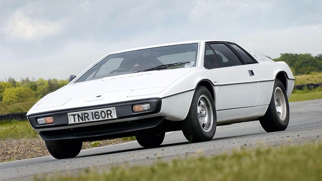 La imagen muestra un Lotus Esprit blanco, un deportivo con un diseño angular característico de los años 70 y 80. Este modelo es conocido por su aparición en la película de James Bond “La espía que me amó”. El vehículo está estacionado en asfalto con césped al fondo, y se destaca por su perfil en forma de cuña y faros escamoteables.