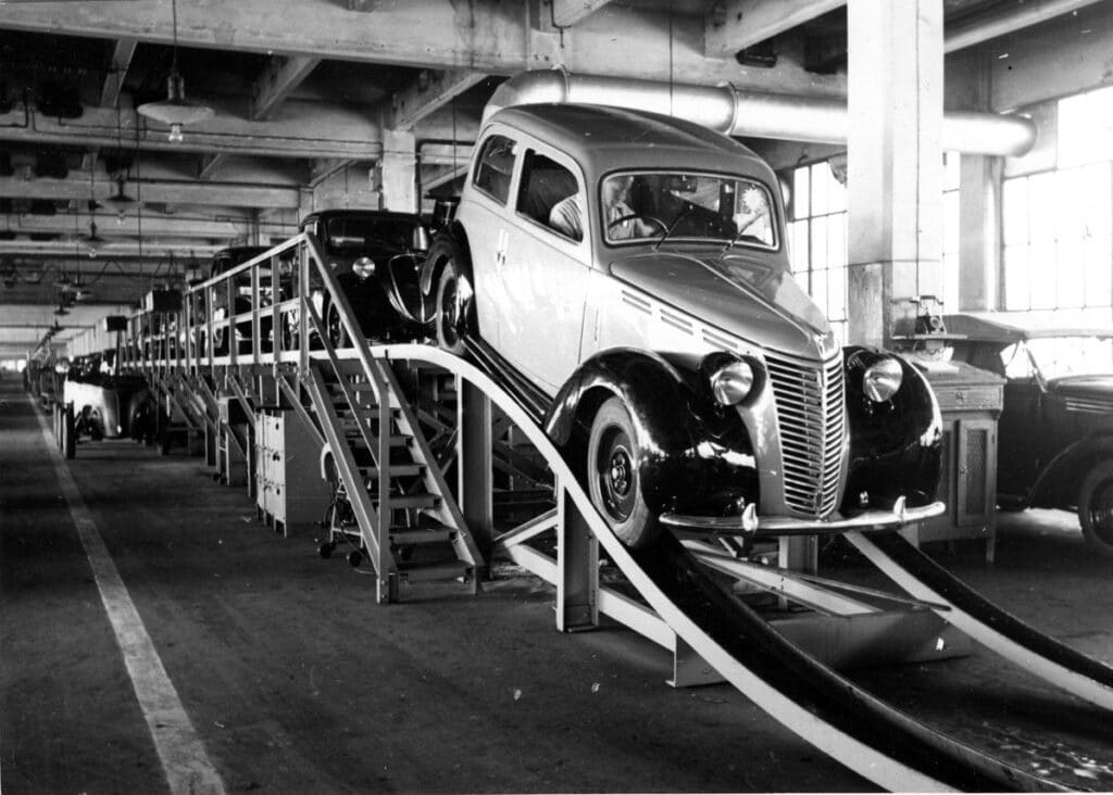 Fotografía en blanco y negro de una línea de ensamblaje en la fábrica de Fiat, probablemente de los años 30. Se ven varios coches en diferentes etapas de finalización en un sistema de cinta transportadora. El coche en primer plano parece completamente ensamblado y tiene un diseño vintage distintivo con una parrilla prominente y una carrocería redondeada. El entorno de la fábrica incluye grandes columnas de soporte y estructuras superiores, típicas de la arquitectura industrial de esa época.