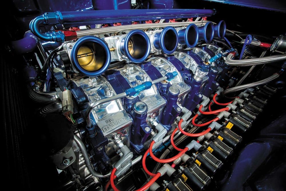 Imagen de un motor de automóvil de alto rendimiento con múltiples tubos y conexiones. Se destacan los colectores de admisión cromados con embudos dorados, cables rojos y tuberías azules sobre un fondo oscuro, lo que sugiere una configuración compleja diseñada para optimizar el rendimiento del motor. Este motor es un Mazda 13B, conocido por su diseño compacto y ligero, con una configuración de dos rotores y una cilindrada total de 1.3 litros (1308 cc).