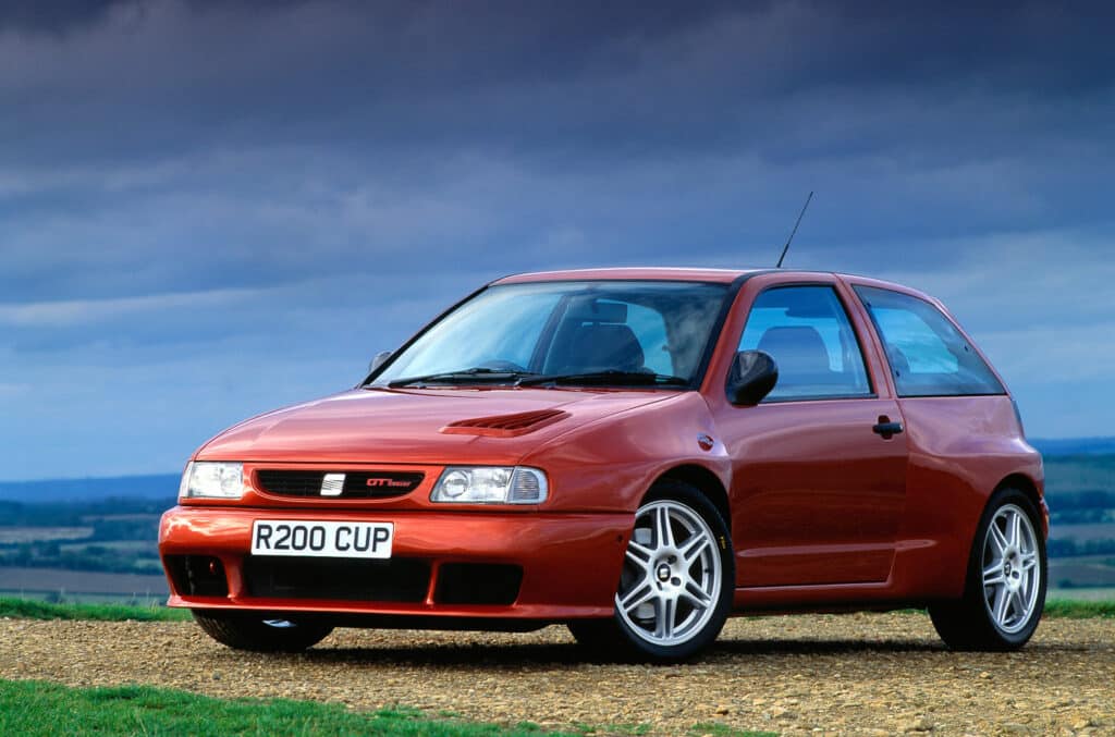 Un coche rojo Ibiza MK2 CUPRA Sport F2 está estacionado sobre una superficie de grava, con un cielo nublado y un horizonte de colinas onduladas en el fondo. El vehículo presenta elementos de diseño deportivo distintivos, como un parachoques delantero con luces antiniebla integradas, una insignia que indica ‘GTI’ en la parrilla y llantas de aleación blancas. La matrícula dice ‘R200 CUP’.