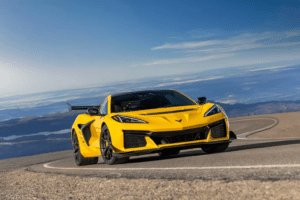 Un Chevrolet Corvette ZR1 amarillo brillante está conduciendo por una carretera sinuosa con un cielo azul claro y montañas en la distancia. El vehículo presenta un diseño agresivo con ángulos pronunciados, grandes tomas de aire y un prominente alerón trasero. La postura dinámica del coche sugiere alto rendimiento y velocidad.