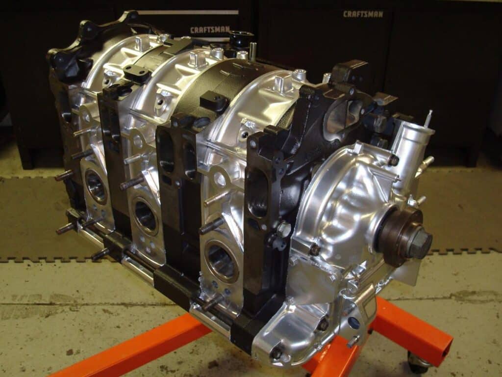 Un motor rotativo 20B, que es un tipo de motor de combustión interna. Este motor es notable por su diseño rotativo único en lugar de los motores de pistón más comunes. El motor parece estar montado en un soporte o estructura naranja, probablemente para exhibición o propósitos de ensamblaje. Tiene un acabado metálico y múltiples puertos y aberturas visibles, indicando dónde se conectarían varios componentes. La marca ‘CRAFTSMAN’ es visible en la herramienta en el fondo, sugiriendo un entorno de taller.