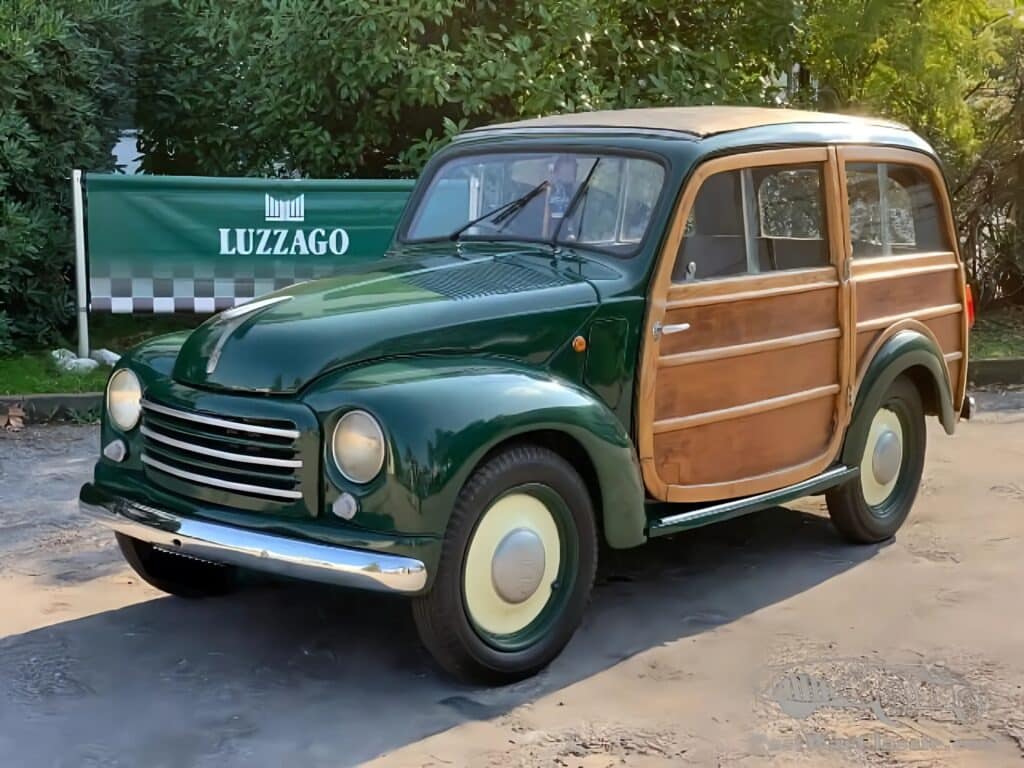 Un Fiat 500 Giardiniera vintage con carrocería de dos tonos y paneles de madera en los laterales. El coche está estacionado sobre una superficie de grava con vegetación al fondo y un cartel que dice ‘LUZZAGO’ detrás, posiblemente indicando la ubicación o concesionario.