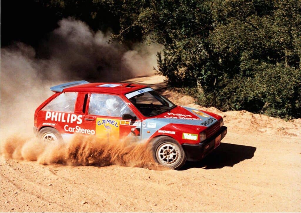 Un coche de rally Seat Ibiza Mk1 Bimotor rojo en acción sobre una pista de tierra, levantando una nube de polvo mientras toma una curva. El vehículo está adornado con logotipos de patrocinadores, incluyendo ‘PHILIPS’ y ‘CAMEL’, indicando su participación en eventos de motorsport competitivo. Este modelo es notable por tener dos motores, uno en cada eje, lo que lo hace único y potente en el mundo de los rallyes.