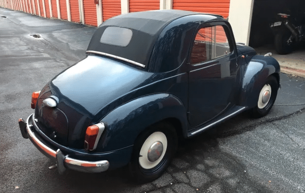 Vista trasera de un Fiat 500 Topolino de 1950, con su característico diseño redondeado y detalles cromados. Este modelo presenta faros integrados en las aletas traseras y una rejilla horizontal en el frente, actualizando su apariencia. El coche está estacionado en un entorno que resalta su estilo clásico y vintage.