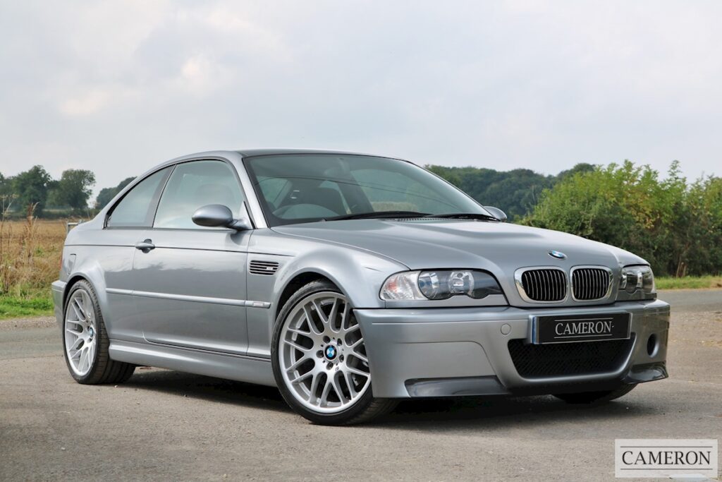 Un BMW M3 E46 Coupe plateado está estacionado al aire libre con un cielo despejado y algo de vegetación en el fondo. El vehículo presenta un diseño distintivo con un parachoques delantero con grandes entradas de aire, rejillas laterales y llantas de aleación de múltiples radios. El nombre “CAMERON” es visible en el área de la placa del automóvil.