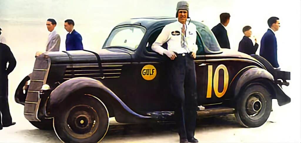 La imagen muestra un coche de carreras vintage con el número 10 y el logotipo de Gulf Oil en su lateral. Junto al coche, hay una persona vestida con una camisa blanca con una insignia y pantalones oscuros que es Bill France, el creador de NASCAR. Detrás de esta persona y del coche, hay varias otras personas con sus rostros también ocultos. La relevancia de esta imagen radica en su importancia histórica para los orígenes de NASCAR y la historia del automovilismo.