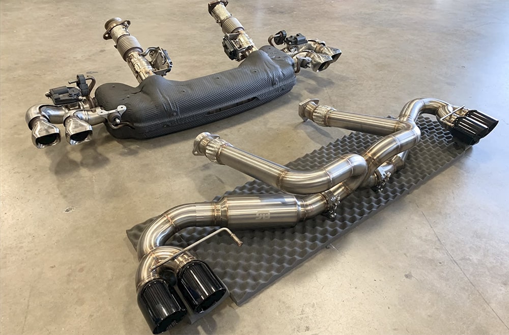 Dos sistemas de escape diferentes del motor del Corvette ZR1, dispuestos sobre un suelo de concreto. El sistema superior es el escape de serie, caracterizado por un diseño más complejo y restrictivo con múltiples cámaras y convertidores catalíticos de fábrica. El sistema inferior es un escape catback, típicamente una modificación del mercado de accesorios diseñada para mejorar el rendimiento al reducir la contrapresión con un diseño más simple y recto, a menudo con menos cámaras y posiblemente sin ciertos dispositivos de control de emisiones como los convertidores catalíticos.