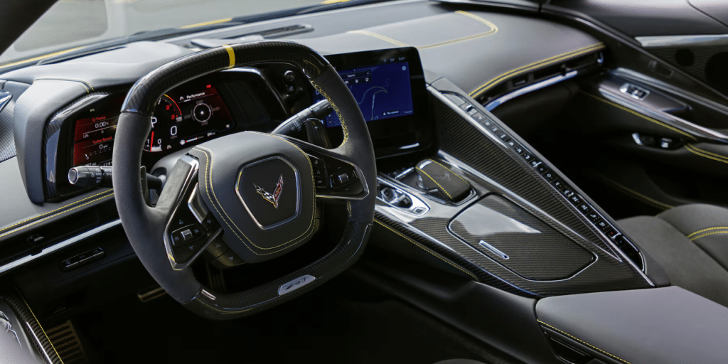 Imagen del interior del Corvette ZR1 2025, enfocada en el área del conductor. El volante tiene el emblema de Chevrolet Corvette. El tablero detrás del volante presenta pantallas digitales con indicadores iluminados en rojo y blanco, incluyendo un tacómetro y posiblemente información de velocidad. En la consola central hay otra pantalla digital grande que muestra contenido de navegación o entretenimiento. El diseño interior incluye texturas de fibra de carbono y materiales de cuero negro con costuras visibles, dando una apariencia lujosa y de alta tecnología.
