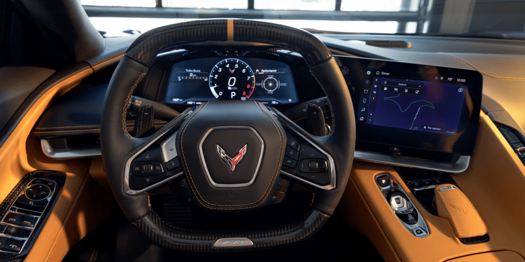 Imagen del interior del Corvette ZR1 2025, enfocada en el volante y el panel de instrumentos. El volante tiene el logotipo de Corvette. El panel de instrumentos incluye un velocímetro digital que marca 0 mph y otros indicadores, mientras que a la derecha hay una pantalla de infoentretenimiento que muestra un mapa. El interior presenta cuero en colores negro y beige.