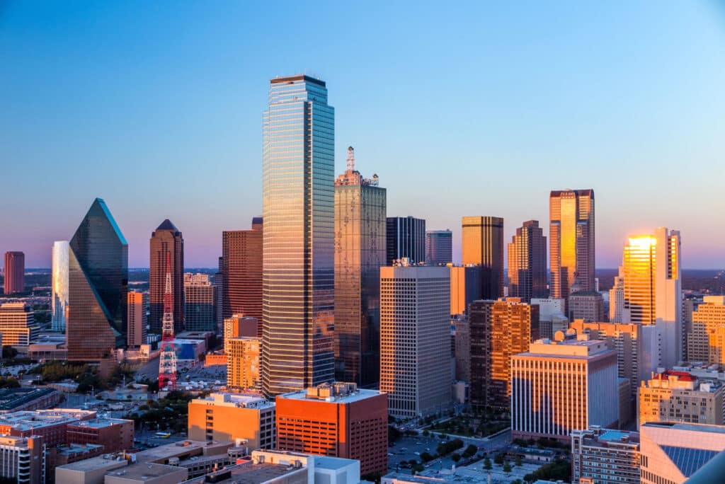 Horizonte de Dallas durante el amanecer o atardecer, con la luz del sol reflejándose en las fachadas de vidrio de varios rascacielos altos. El cielo está despejado con un gradiente de naranja cálido cerca del horizonte a un azul más profundo más arriba. Esta ciudad fue la sede de la ZCON en 1988.