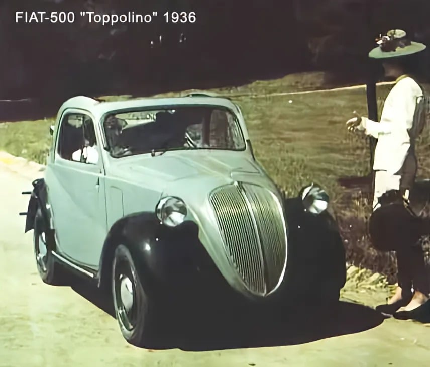 Un Fiat 500 Topolino vintage de 1936 estacionado al borde de la carretera con césped y árboles al fondo. El coche tiene una carrocería redondeada, una parrilla frontal prominente y faros distintivos. Al lado del coche, hay una persona vestida de blanco sosteniendo algo que parece una bandeja o plato. Detrás de esta persona, otra figura parece estar interactuando con un animal, posiblemente alimentándolo.