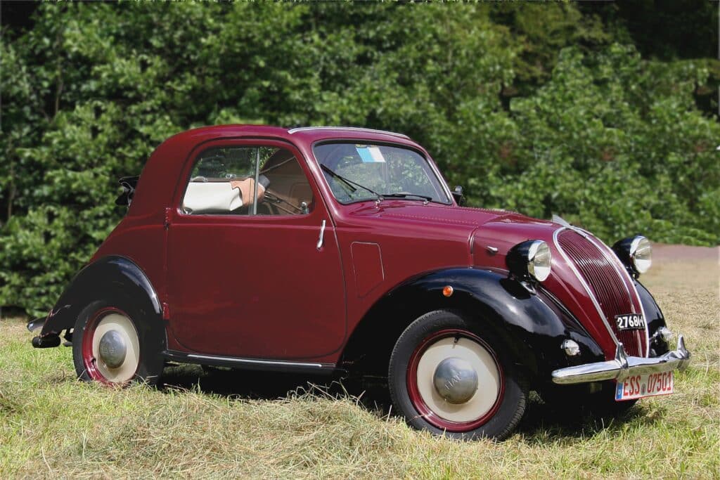Un clásico Fiat 500 Topolino de color rojo oscuro con una sección frontal negra, estacionado sobre césped con árboles al fondo. El coche tiene neumáticos con paredes blancas y un techo solar abierto a través del cual se ve parte del brazo de una persona. La matrícula es visible en la parte delantera del coche.
