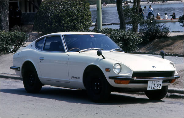 Imagen de un automóvil deportivo clásico Fairlady Z de color blanco, estacionado al aire libre. El vehículo tiene un diseño coupé de dos puertas con faros redondos y una línea larga y baja que se extiende desde el frente hasta la parte trasera. En el fondo, hay árboles y personas disfrutando del entorno en lo que parece ser un parque o área recreativa. La placa del vehículo muestra caracteres alfanuméricos que indican su registro.