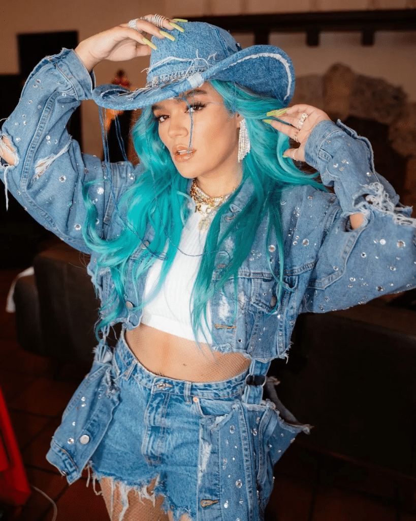Karol G viste un conjunto de mezclilla con chaqueta y shorts, con el cabello largo de color teal, grandes aretes de aro y uñas pintadas de varios colores. El conjunto de mezclilla está adornado con lo que parecen ser incrustaciones de pedrería.