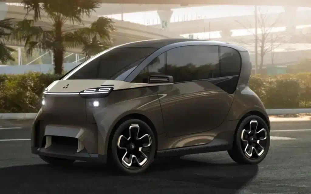 Un Liux Geko, un vehículo eléctrico compacto y moderno en tonos de gris y negro, está estacionado al aire libre. Presenta un parabrisas grande y ventanas laterales, ruedas negras distintivas con patrones blancos, rodeado de árboles y parte de un edificio desenfocado en el fondo.