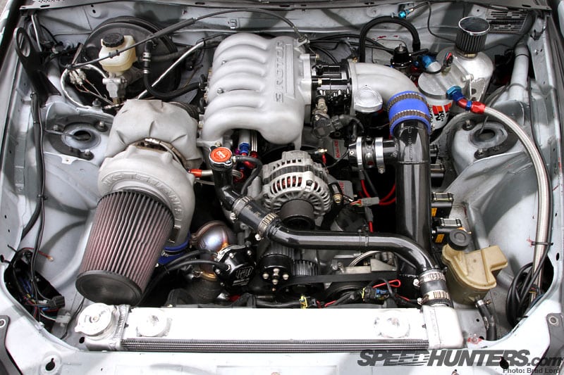 La imagen muestra una vista de cerca del compartimento del motor, destacando un motor rotativo 20B modificado de Mad Mike. Este tipo de motor es notable por su diseño rotativo único en lugar de los motores de pistón más comunes. El motor parece estar fuertemente personalizado con piezas de posventa, lo que podría indicar que está construido para alto rendimiento o propósitos de carreras. La presencia de varios componentes como el sistema de admisión, el sistema de enfriamiento y posiblemente elementos de inducción forzada como turbocompresores lo hacen interesante para los entusiastas de los automóviles o aquellos interesados en la ingeniería mecánica y las modificaciones automotrices.