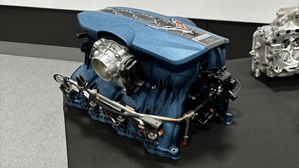 Araña de admisión del motor del Corvette ZR1. Este componente, pintado de azul, es parte del sistema de admisión de aire que distribuye el aire a cada cilindro del motor. La imagen muestra detalles intrincados como tubos, conectores y un cuerpo de mariposa en el centro superior. La araña de admisión es crucial para el rendimiento del motor, asegurando una distribución uniforme del aire.