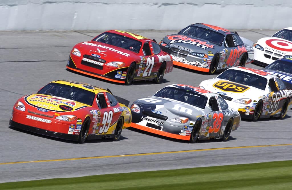 La imagen muestra un grupo de coches de la NASCAR compitiendo de cerca en una pista de asfalto. Los coches están pintados de colores brillantes con varios logotipos de patrocinadores y números visibles. El coche líder es rojo con el número 20, seguido por otro coche rojo con el número 99, y varios otros en proximidad, incluyendo un coche negro y naranja con el número 31, un coche blanco y azul con el número 48, y un coche amarillo con el número 38. Esta imagen captura la intensidad y competitividad de una carrera de NASCAR, destacando las altas velocidades y la cercanía entre los vehículos.