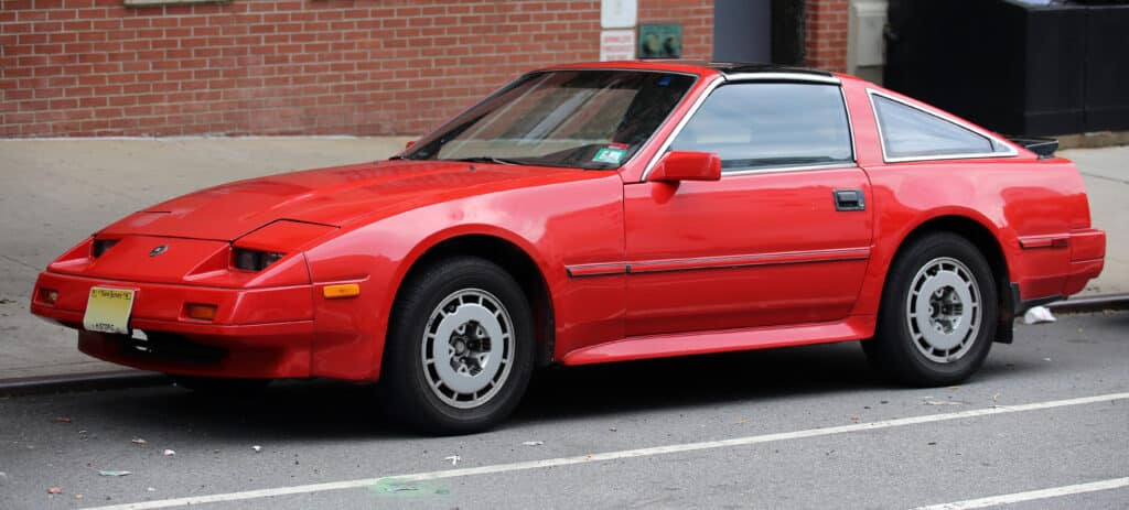 Imagen de un Fairlady Z Z31 rojo, un clásico deportivo de Nissan, estacionado en la calle. Características distintivas como faros emergentes y techo T-top.