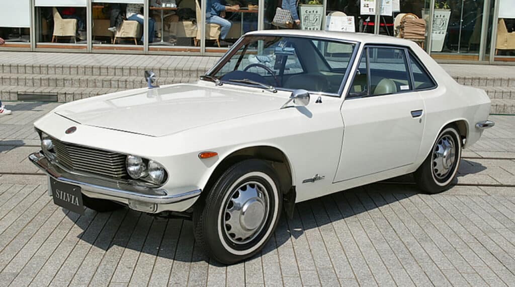 Nissan Silvia de 1964 en color blanco, estacionado al aire libre. El vehículo presenta un diseño clásico distintivo con un capó largo, faros redondeados y parachoques cromados. Las líneas elegantes y la estética vintage del coche lo hacen interesante y relevante para los entusiastas de los automóviles clásicos y la historia automotriz.