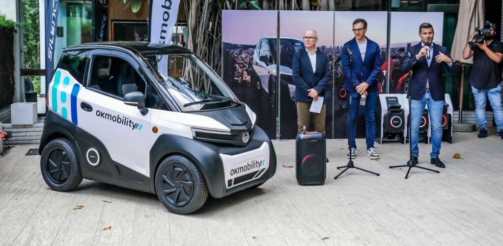 Un pequeño vehículo eléctrico blanco con el logo “okmobility” en el lateral está estacionado frente a un fondo con imágenes de personas y vehículos. Tres personas están de pie junto al vehículo.
