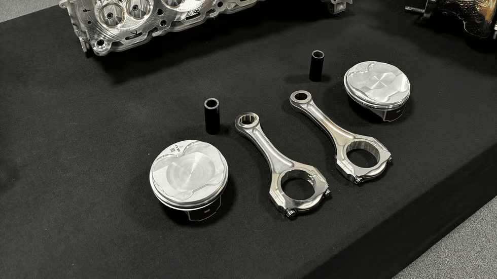 Dos pistones y dos bielas del motor LT7 del Corvette ZR1, exhibidos sobre una superficie negra. Los pistones tienen un acabado metálico con rebajes para las válvulas en la corona, indicando su diseño para aplicaciones de alto rendimiento. Las bielas también son metálicas y muestran una construcción robusta con una sección de viga grande para mayor resistencia. Los pistones son supercuadrados, con un diámetro de 104.25 mm y una carrera de 85.8 mm, lo que permite mayores revoluciones por minuto (RPM) y mejora la eficiencia volumétrica. Esta imagen destaca los componentes internos cruciales para el funcionamiento y rendimiento del motor LT7.