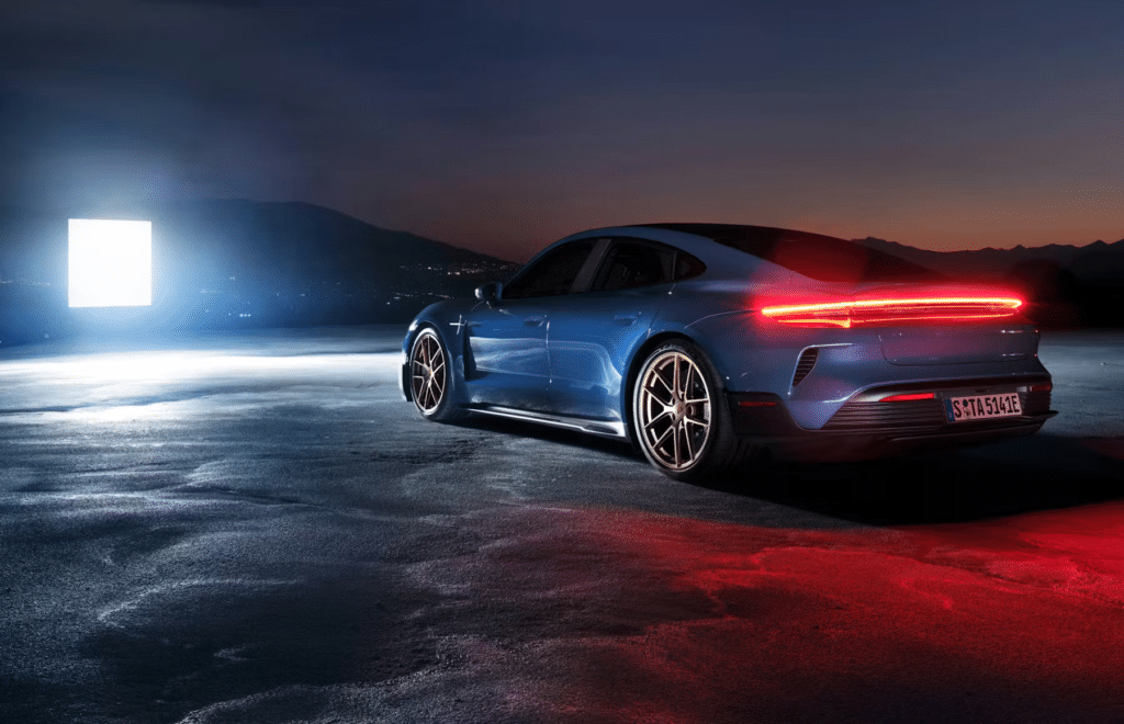 Un Porsche Taycan azul eléctrico estacionado sobre una superficie mojada, reflejando las luces del coche. El vehículo está en un ángulo de tres cuartos trasero, mostrando su diseño elegante y la distintiva tira de luz trasera. Al fondo, se ve un paisaje montañoso tenuemente iluminado bajo un cielo crepuscular, con una fuente de luz cuadrada artificial a la izquierda, creando una escena dramática y de alto contraste.