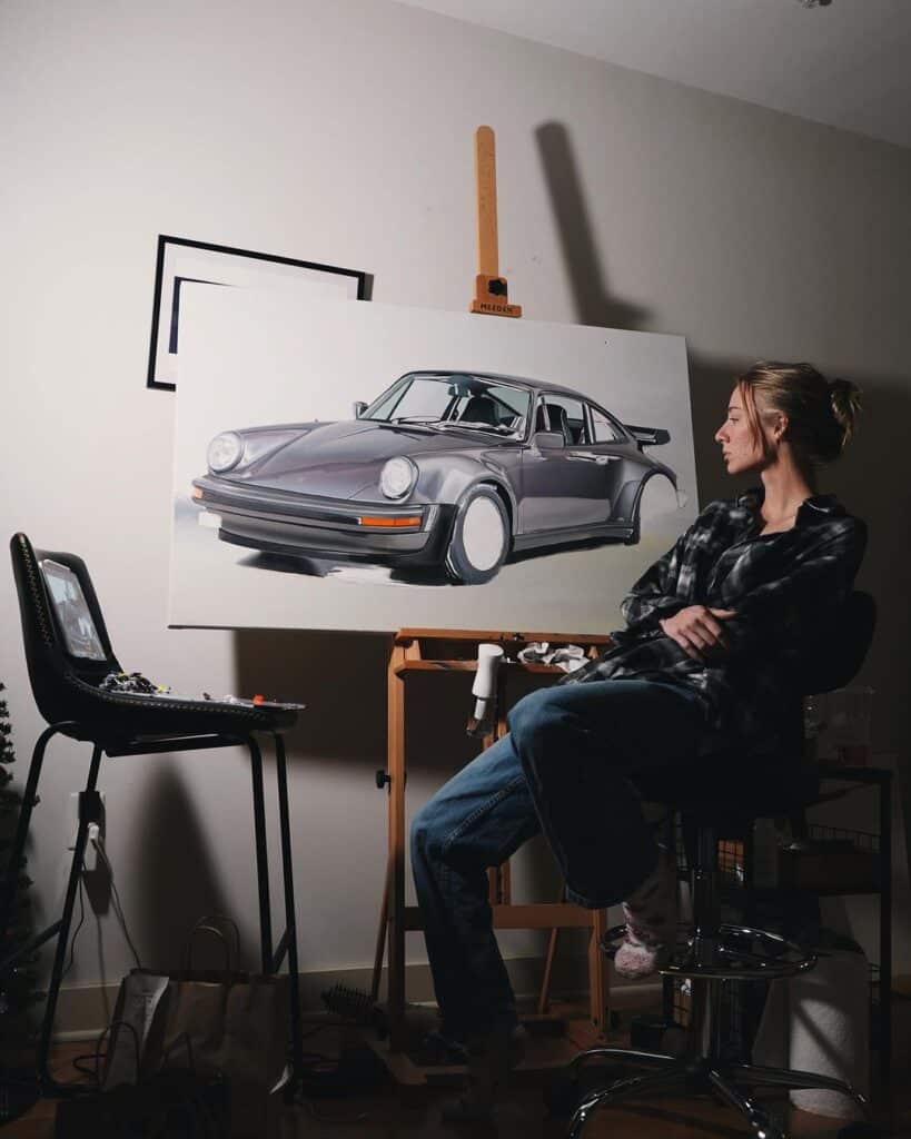 La imagen muestra a Rae Roberts sentada en un taburete de estudio frente a un gran lienzo sobre un caballete. En el lienzo hay una pintura detallada de un automóvil Porsche 911 clásico en tonos grises y negros. Rae, que está de perfil y no se le distingue la cara, viste una chaqueta de cuero negra y parece estar contemplando la obra o tomando un descanso. A su lado hay una silla con paletas de pintura y pinceles, lo que indica que se encuentra en un espacio dedicado a la pintura o arte. La obra es de la artista Rae Roberts.