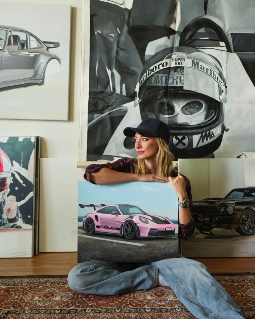 La imagen muestra a Rae Roberts posando junto a varios de sus cuadros. Los cuadros presentan detalladas pinturas de automóviles Porsche clásicos. Rae está de pie en un estudio de arte, rodeada de sus obras, lo que resalta su dedicación y talento en la pintura automotriz.