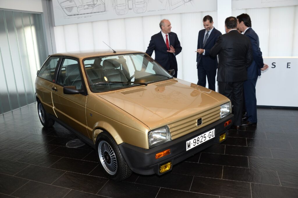 Un coche SEAT Ibiza dorado está exhibido en un entorno interior, posiblemente un showroom o espacio de exposición. La matrícula del coche es ‘M-9825-GU’. Este vehículo fue el primer coche del Rey Felipe VI de España, un regalo de su padre, el Rey Juan Carlos I, en su 18 cumpleaños en 1986. El coche tiene detalles exclusivos como un volante de Porsche y asientos Recaro.