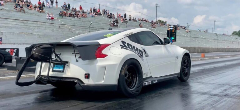 Un coche deportivo blanco, específicamente un Nissan 370Z, en la línea de salida de una pista de carreras de aceleración durante el evento ZCON. El vehículo está equipado con un alerón trasero y neumáticos de carreras, indicando modificaciones para una aceleración de alto rendimiento. El coche está listo para lanzarse por la pista, con las luces del ‘árbol de Navidad’ visibles en el fondo, indicando el inicio de una carrera. La presencia de espectadores en las gradas añade a la atmósfera competitiva del evento.
