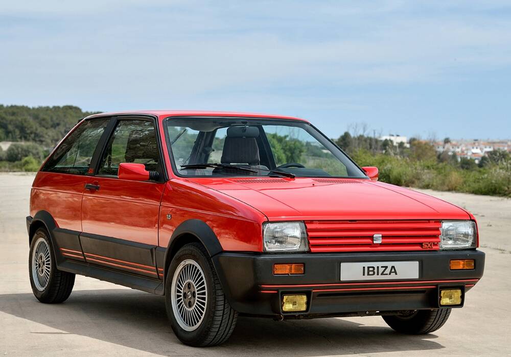 Un coche hatchback rojo de cuatro puertas, modelo SEAT Ibiza SXi de 1990, está estacionado al aire libre sobre una superficie pavimentada, con vegetación y un cielo despejado de fondo. El nombre del modelo ‘IBIZA’ es visible en la parte frontal del vehículo.