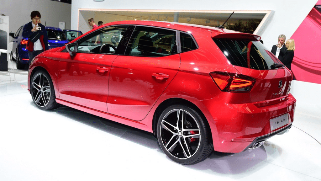 Un coche SEAT Ibiza rojo de 2017 está exhibido en un salón del automóvil. El vehículo está posicionado para mostrar su perfil, destacando su diseño elegante y llantas deportivas de aleación. El entorno incluye una iluminación brillante y un suelo blanco, lo que contrasta con el color vibrante del coche. Hay personas en el fondo, indicando la naturaleza pública del evento.