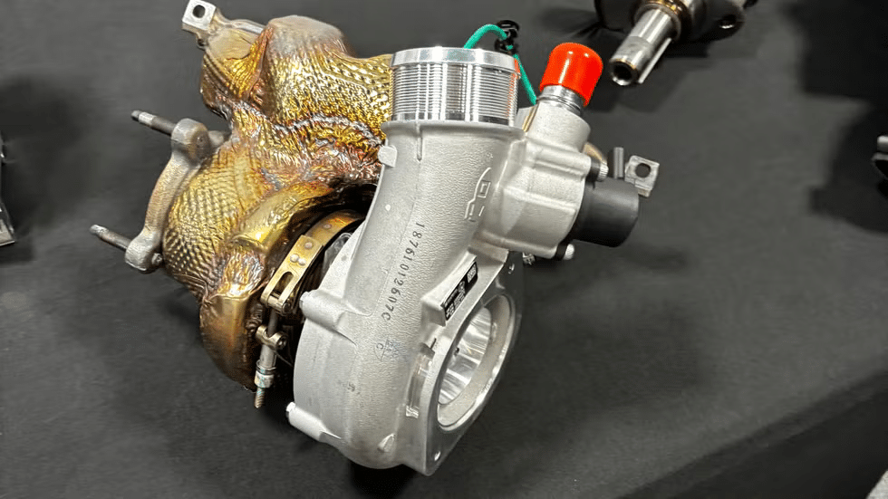 Turbo del motor del Corvette ZR1 exhibido sobre una superficie negra. El turbo tiene un acabado plateado prominente con algunas áreas envueltas en papel de oro resistente al calor. Hay varios tubos y conectores adjuntos, uno con una tapa roja y otro con una tapa verde. El diseño intrincado y los componentes muestran la ingeniería avanzada de las piezas automotrices de alto rendimiento.