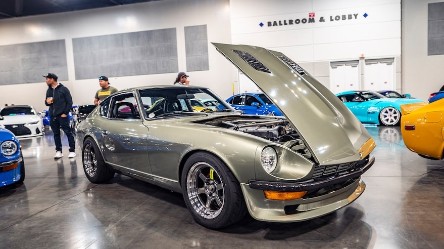 Un Fairlady Z clásico con el capó abierto, exhibido en la ZCON. El coche, de estilo vintage, está pintado en un color metálico y tiene molduras y ruedas negras. Al fondo, se pueden ver otros coches y asistentes caminando, indicando que es parte de una exposición de automóviles.