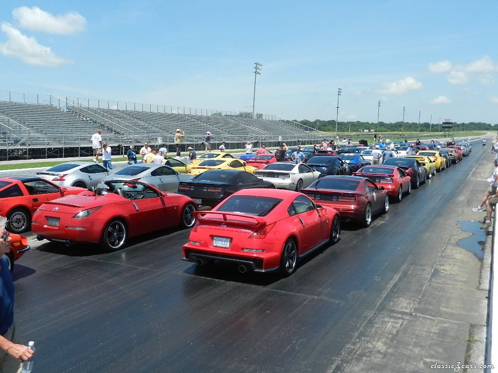 Una fila de varios coches deportivos en una pista de carreras durante la ZCON. Los coches son de diferentes modelos y colores, predominantemente rojos y amarillos, con algunos negros y blancos. Están posicionados uno al lado del otro, preparados para una carrera o exhibición. Al fondo, se puede ver un cielo despejado y una tribuna con espectadores.