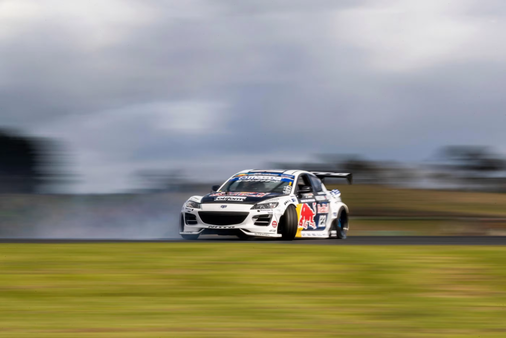 La imagen muestra un coche de carreras Mazda RX-8 con motor rotativo en movimiento en una pista. El coche es predominantemente blanco con logotipos de patrocinadores, incluyendo el prominente logotipo de Red Bull en el lateral. El fondo está borroso debido a la alta velocidad del vehículo, enfatizando su movimiento y velocidad, lo que lo hace interesante al capturar un momento dinámico en el automovilismo.