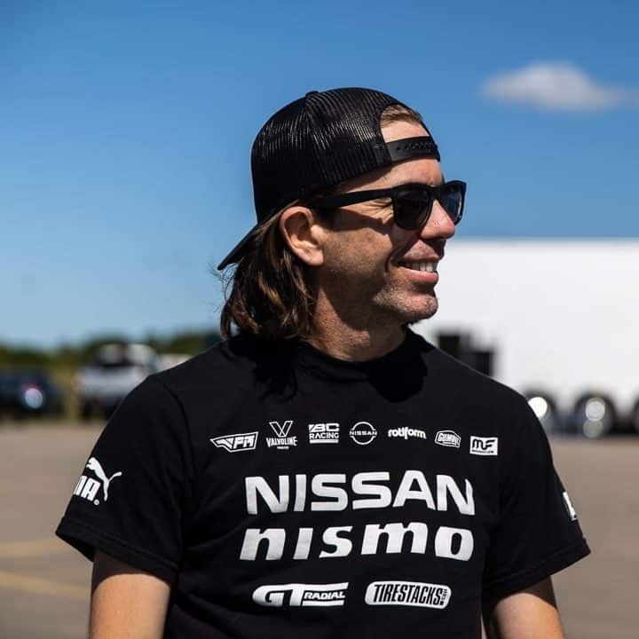 Bryan Heitkotter con una camiseta negra con múltiples logotipos y el texto ‘NISSAN NISMO’ en el pecho, en la ZCON. El fondo muestra un área exterior con cielo despejado.