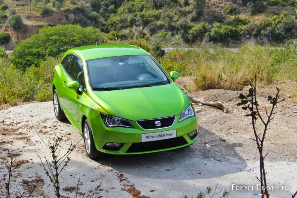 Un coche SEAT Ibiza verde brillante de 2008 está estacionado en un camino de tierra irregular con vegetación y colinas al fondo. El coche está posicionado en un ángulo que muestra su perfil frontal y lateral. La iluminación sugiere que es de día. El diseño del coche incluye faros delanteros estilizados y llantas de aleación deportivas.