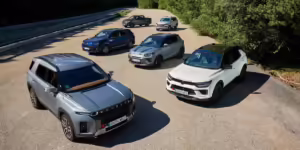 Una imagen de cinco SUVs de la marca KGM (anteriormente SsangYong) estacionados en formación en una carretera rodeada de árboles. Los vehículos varían en color, incluyendo tonos de azul, negro, gris y blanco. La luz del sol ilumina la escena, destacando los diseños modernos y robustos de los SUVs.